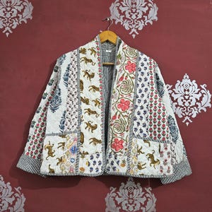 Peut inclure: Veste matelassée avec un motif patchwork de tigres, de fleurs et de motifs géométriques. La veste est blanche avec des touches de rouge, de bleu et de noir. Suspendue à un cintre en bois.