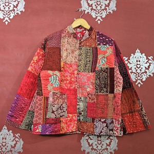 Peut inclure: Veste patchwork aux tons rouges, roses et bordeaux, suspendue à un cintre en bois. La veste présente une variété de carrés de tissu à motifs, créant un design unique et coloré. La veste a un col mandarin et des fermetures à boutons.