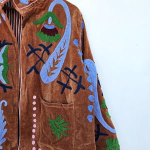 May include: A brown velvet jacket with detailed embroidery in blue, green, and pink. It has a high collar, front pockets, and a striped lining. The jacket is decorated with floral and abstract patterns.