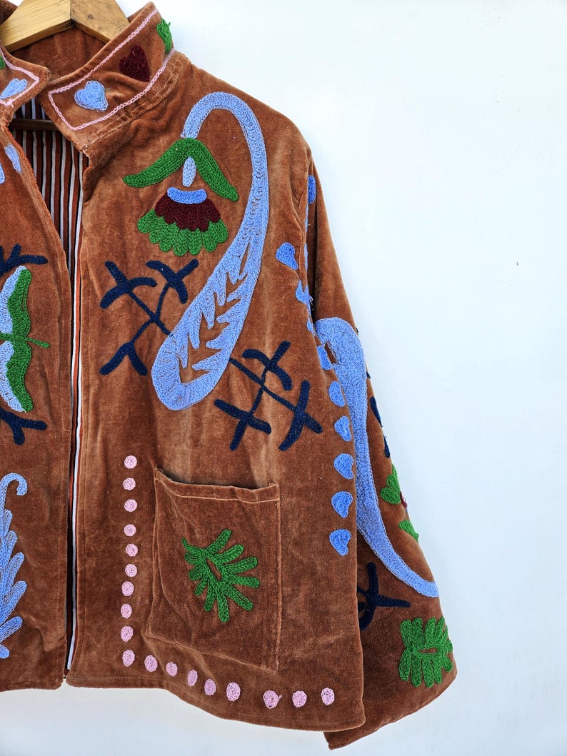 May include: A brown velvet jacket with detailed embroidery in blue, green, and pink. It has a high collar, front pockets, and a striped lining. The jacket is decorated with floral and abstract patterns.