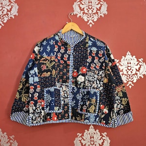 May include: A quilted jacket with a patchwork design featuring floral patterns in red, blue, and green. The jacket has a stand-up collar, two pockets, and striped trim. It is hanging on a wooden hanger against a red background.