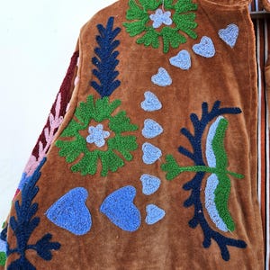 May include: Brown velvet jacket with colourful embroidered floral and heart designs. The jacket features green, blue, and pink embroidery, with a striped lining visible at the collar. The jacket has a pocket on the lower front.