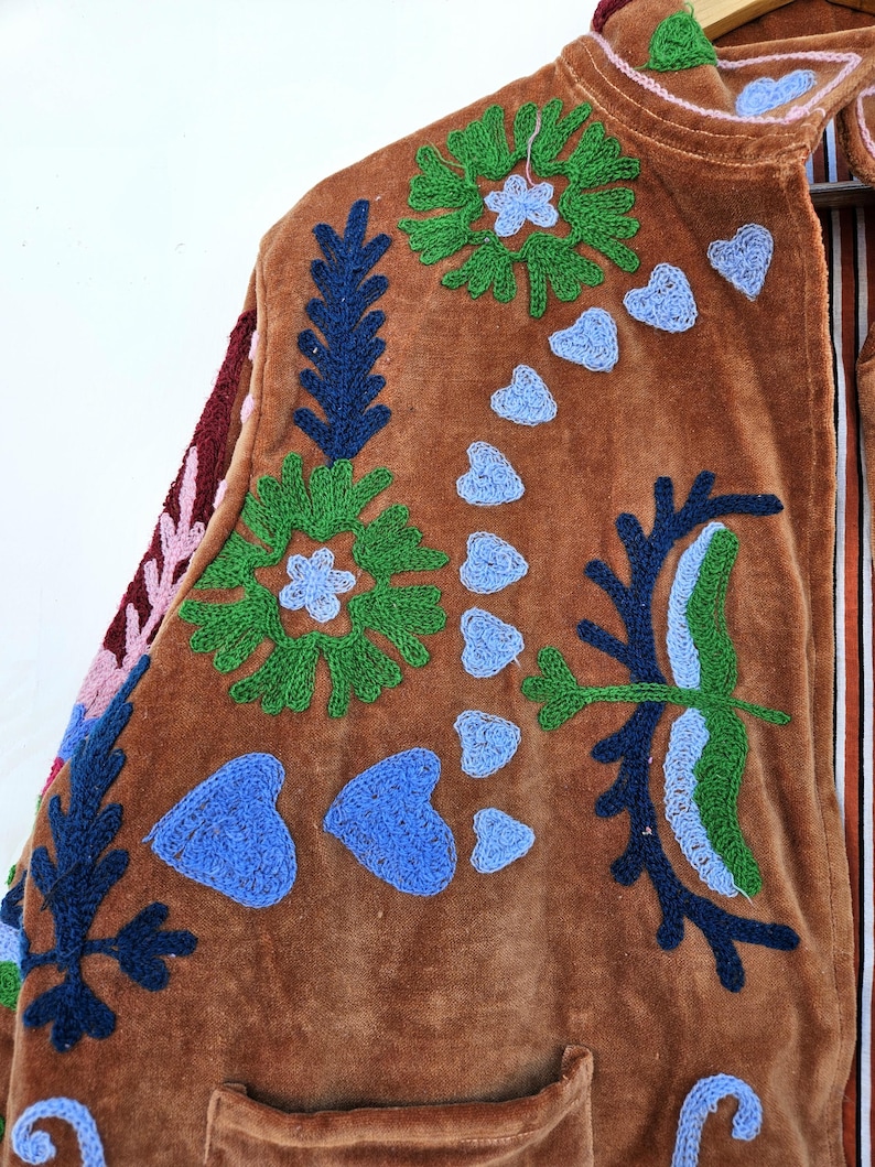 May include: Brown velvet jacket with colourful embroidered floral and heart designs. The jacket features green, blue, and pink embroidery, with a striped lining visible at the collar. The jacket has a pocket on the lower front.