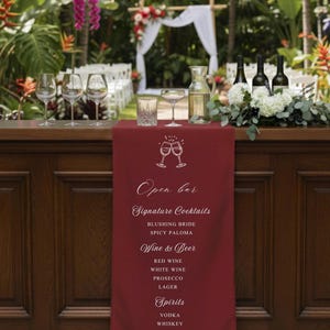 May include: A burgundy open bar sign listing cocktails, wine, beer, spirits, and soft drinks. The sign is on a wooden bar, with glasses and bottles, set against a garden backdrop with a wedding arch.