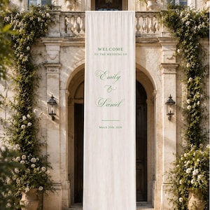 May include: A long, vertical off-white fabric banner hanging in front of a stone building entrance. The banner features dark green text that reads: WELCOME TO THE WEDDING OF Emily & Daniel, followed by a horizontal line and the date March 25th, 2026.