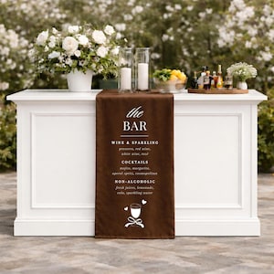 May include: A white bar with a brown runner featuring the text "the BAR" and a list of drinks. The bar is decorated with flowers, candles, and a tray of bottles. The bar is set up outdoors.