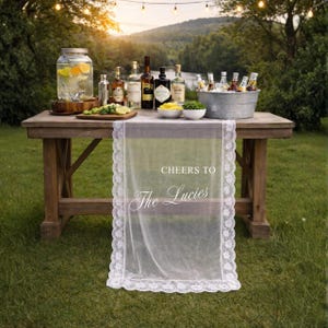 May include: A rustic wooden table set outdoors with various bottles of liquor, a large glass beverage dispenser, and a metal ice bucket filled with drinks. A sheer white lace runner with the text "Cheers to The Lucies" is draped over the table.