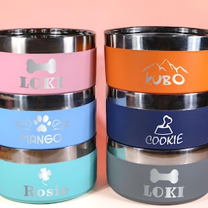 May include: A stack of six stainless steel pet bowls in various colors: pink, blue, teal, orange, navy, and gray. Each bowl has a silver rim and a name printed on it, along with a graphic such as a paw print or bone.
