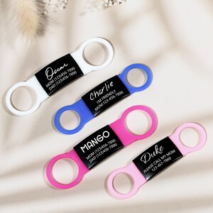 Op de afbeelding: Vier kleurrijke dierenriemhouders in wit, blauw, roze en lichtroze. Elke tag heeft een zwart label met de naam en contactgegevens van het huisdier. De tags zijn ontworpen om aan de halsband van een huisdier te bevestigen.