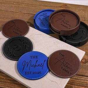 Op de afbeelding: Een verzameling gepersonaliseerde leren onderzetters in verschillende kleuren, waaronder bruin, blauw en zwart. Elke onderzetter heeft aangepaste tekst, zoals familienamen en data, met witte stiksels rond de randen. De onderzetters zijn gerangschikt op een lichtgekleurd oppervlak.