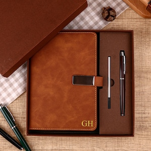 May include: A brown leather journal with a silver clasp and the initials "GH" in gold, presented in a matching gift box. Includes a brown pen and a silver pen refill. The set is displayed on a wooden surface.