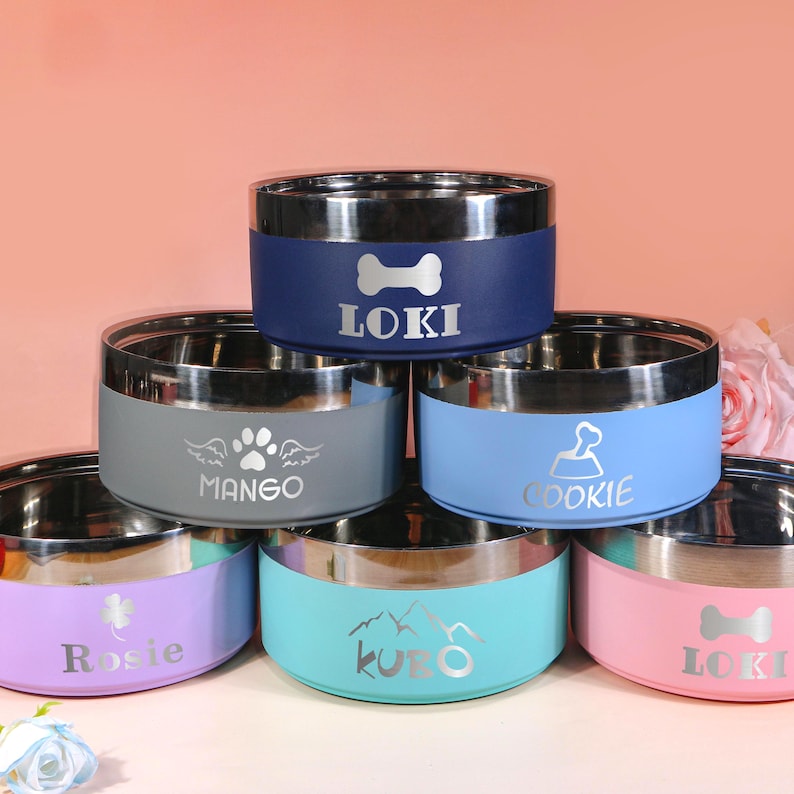 May include: A collection of stainless steel pet bowls in various colors, including blue, gray, light blue, pink, purple, and teal. Each bowl features a silver rim and a personalized name or design, such as a bone, paw prints, or mountains. The bowls are designed for pets.