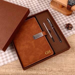 May include: A brown leather journal with a metal clasp, presented in a matching gift box. The journal is embossed with the initials "GH" in gold. A pen and stylus are included in the box, creating a complete writing set.