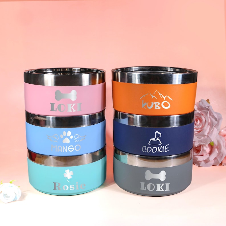 May include: A stack of six stainless steel pet bowls in various colors: pink, blue, teal, orange, navy, and gray. Each bowl has a silver rim and a name printed on it, along with a graphic such as a paw print or bone.