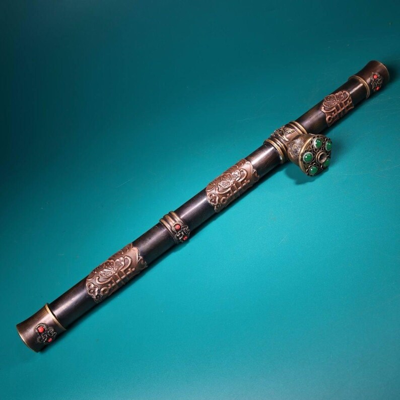 May include: A long, dark pipe with ornate metalwork and decorative accents. The pipe features intricate patterns, red and green gemstones, and a detailed design. The overall aesthetic suggests a vintage or antique style, with a focus on craftsmanship and detail.
