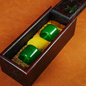 May include: Two cylindrical, vibrant green jade rings rest inside a dark wooden box with a hinged lid. The rings sit on a yellow satin-like fabric. The box is open, revealing the contents. The lid is decorated with green accents.