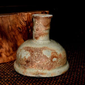 Peut inclure: Petite bouteille en céramique de style antique, avec une base arrondie et un col étroit. La bouteille présente une teinte vert clair avec des taches marron et brun rougeâtre, lui donnant un aspect vieilli. Il s'agit probablement d'un objet décoratif.