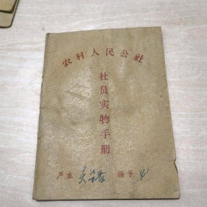 May include: A vintage booklet with Chinese characters in red ink on a tan cover. The text includes "Rural People's Commune" and "Member's Handbook". Handwritten characters and a number are visible at the bottom.