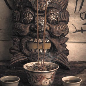 May include: A detailed image of a wooden carved mask with a fierce expression, incense sticks, a floral patterned bowl, and two small teacups. Smoke rises from the incense, creating a spiritual atmosphere. The scene is set on a dark, textured surface.