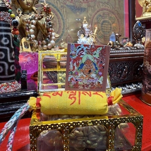 Könnte beinhalten: Ein gelbes Stoffamulett mit roter tibetischer Schrift, das mit einer roten Schnur gebunden ist und auf einer goldfarbenen Metallschachtel mit kleinen goldenen Objekten liegt. Dahinter steht eine Karte mit einer bunten Gottheitsdarstellung.