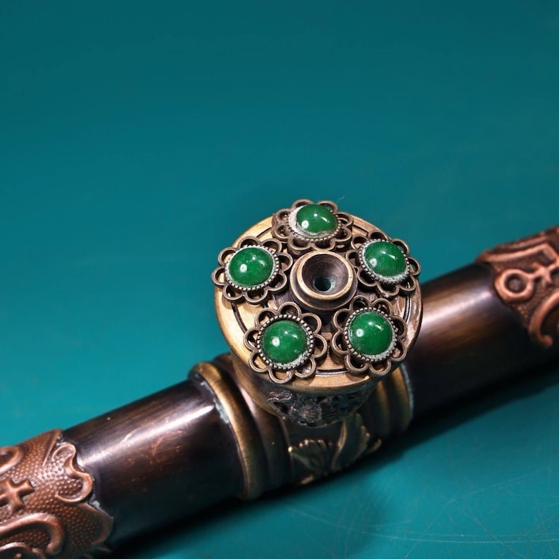 May include: An antique smoking pipe with a bronze finish. The pipe's bowl is adorned with five round, green gemstones set in a decorative, floral-patterned metal casing. The stem is dark brown with ornate metal detailing.