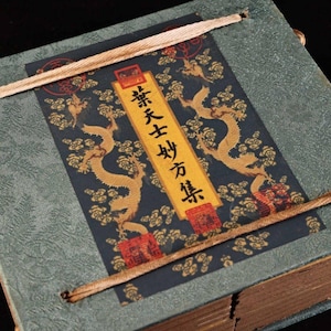 Könnte beinhalten: Ein antikes Buch mit tealfarbenem Einband und einer dekorativen Tafel mit goldenen Drachen und Wolken. Das Panel hat chinesische Schriftzeichen in vertikaler Form. Eine beige Schnur wird verwendet, um das Buch zu binden.