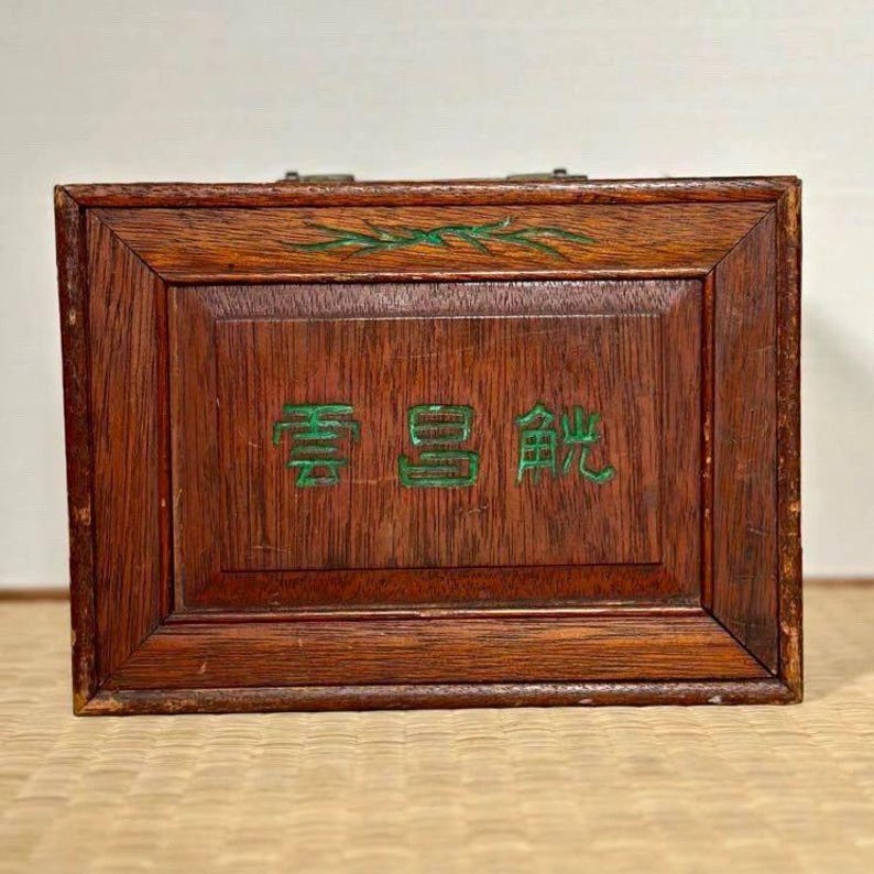 May include: A rectangular wooden box with a dark brown finish. The box features a framed design with a central panel that has green Chinese characters. A green leaf design is at the top. The box has a metal clasp.