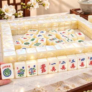 Puede incluir: Un conjunto de fichas de Mahjong de color blanco roto nacarado dispuestas en formación rectangular. Las fichas presentan caracteres, símbolos y números grabados en tinta roja, azul y verde. Una ficha sostenida en primer plano muestra la palabra JOKER.