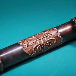 May include: Close-up of a dark brown object with a decorative copper-colored band. The band features an intricate pattern of geometric shapes and floral designs. The object appears to be a handle or part of a larger item.