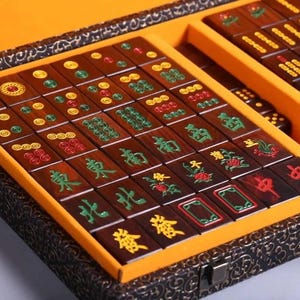 May include: A wooden Mahjong set with brown tiles featuring green, red, and yellow symbols and characters. The tiles are arranged in a yellow-lined box with a decorative, dark-colored exterior. The set includes dice.