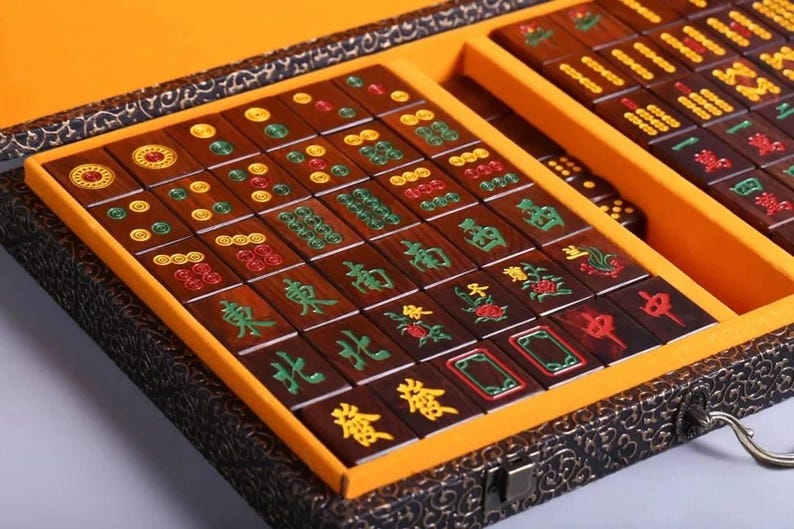May include: A wooden Mahjong set with brown tiles featuring green, red, and yellow symbols and characters. The tiles are arranged in a yellow-lined box with a decorative, dark-colored exterior. The set includes dice.