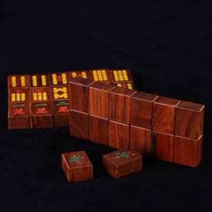 May include: Wooden Mahjong tiles in a variety of colors, including red, green, and yellow. The tiles are arranged in stacks and rows, with some individual tiles visible. The game pieces are made of a dark wood with a rich grain.