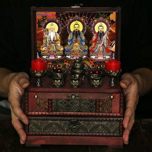 Può includere: Un cofanetto in legno scuro con coperchio a cerniera, raffigurante l'immagine di tre figure con aureole. La scatola ha un cassetto ed è decorata con elaborate decorazioni in metallo. Contiene piccole tazze di metallo, un bruciatore di incenso e due candele rosse.