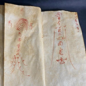 May include: Aged paper with red ink calligraphy. The script features stylized characters and symbols, including a spiral and what appears to be a figure. The paper has a worn, vintage appearance, suggesting age and historical significance.