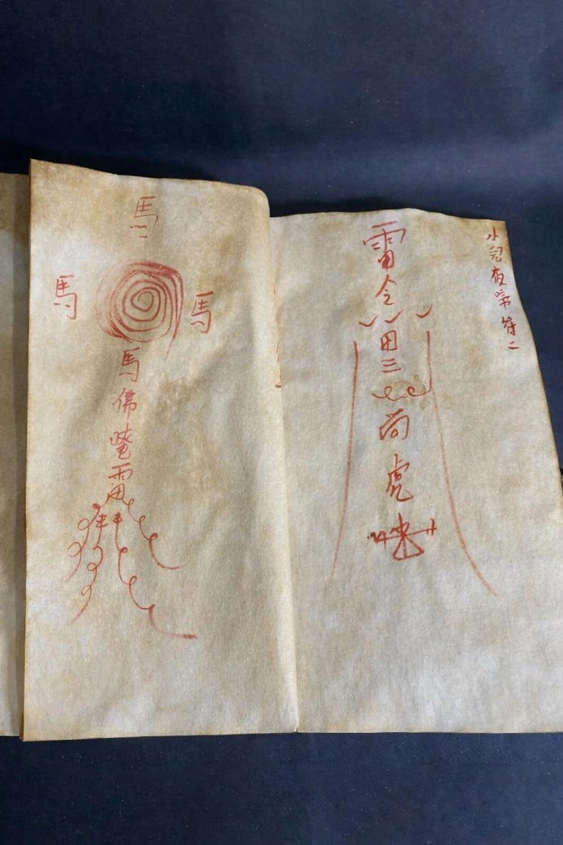 May include: Aged paper with red ink calligraphy. The script features stylized characters and symbols, including a spiral and what appears to be a figure. The paper has a worn, vintage appearance, suggesting age and historical significance.