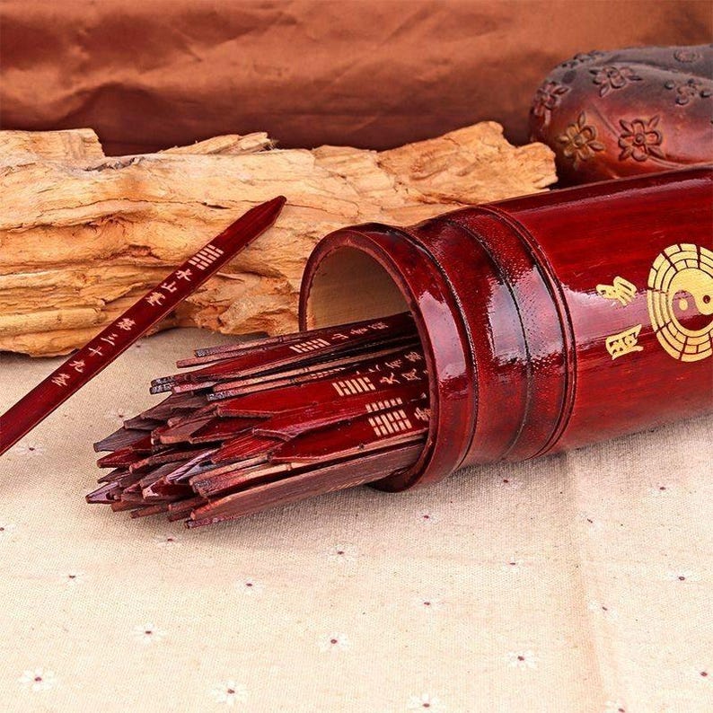 May include: A red wooden fortune telling set with a cylindrical container and multiple sticks. The container has a gold Yin Yang symbol and Chinese characters. A single stick lies outside the container, also with Chinese characters.