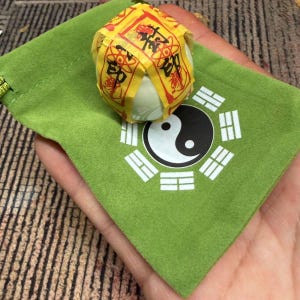 Puede incluir: Un pequeño objeto redondo y blanco envuelto en papel amarillo con caracteres chinos rojos y negros, descansando sobre una bolsa de terciopelo verde. La bolsa presenta un símbolo de Yin y Yang blanco rodeado de ocho trigramas.