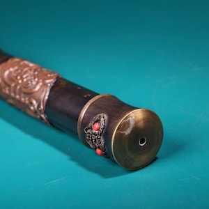 May include: Close-up of a decorative sword handle. The handle features ornate bronze-colored metalwork with a dark wooden grip. Red stone accents are visible on the end cap. The sword handle is set against a teal background.