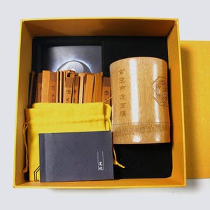May include: A boxed set featuring a bamboo cup with etched text and a geometric design, a stack of bamboo sticks, a yellow drawstring bag, and a small black notebook. The box is yellow with a black interior.