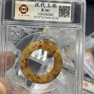 Può includere: Anello in giada antica, di forma circolare, presentato in un contenitore di plastica trasparente. L'anello è di colore beige chiaro con macchie marrone rossiccio. Il contenitore ha un'etichetta con caratteri cinesi. Misure: 35,2 mm x 4,7 mm, peso: 6,2 g.
