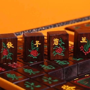 May include: Close-up of four wooden Mahjong tiles with Chinese characters and floral designs in red, green, and gold. The tiles are arranged on a dark brown game board within a yellow case.
