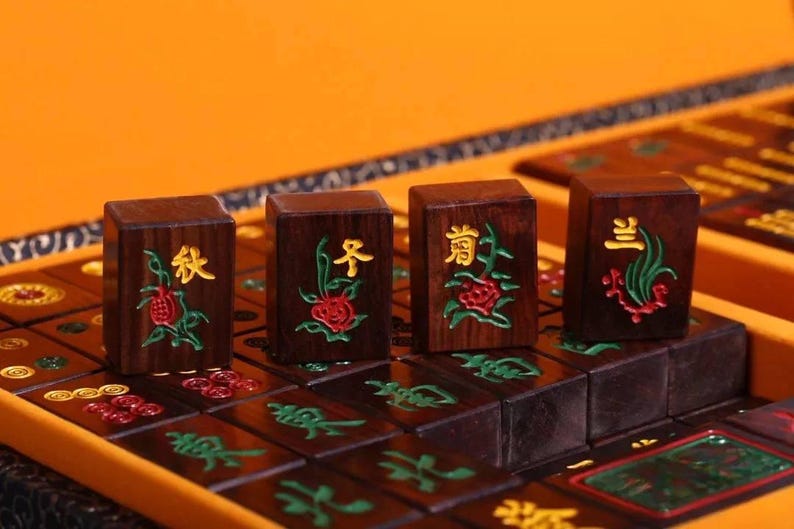 May include: Close-up of four wooden Mahjong tiles with Chinese characters and floral designs in red, green, and gold. The tiles are arranged on a dark brown game board within a yellow case.