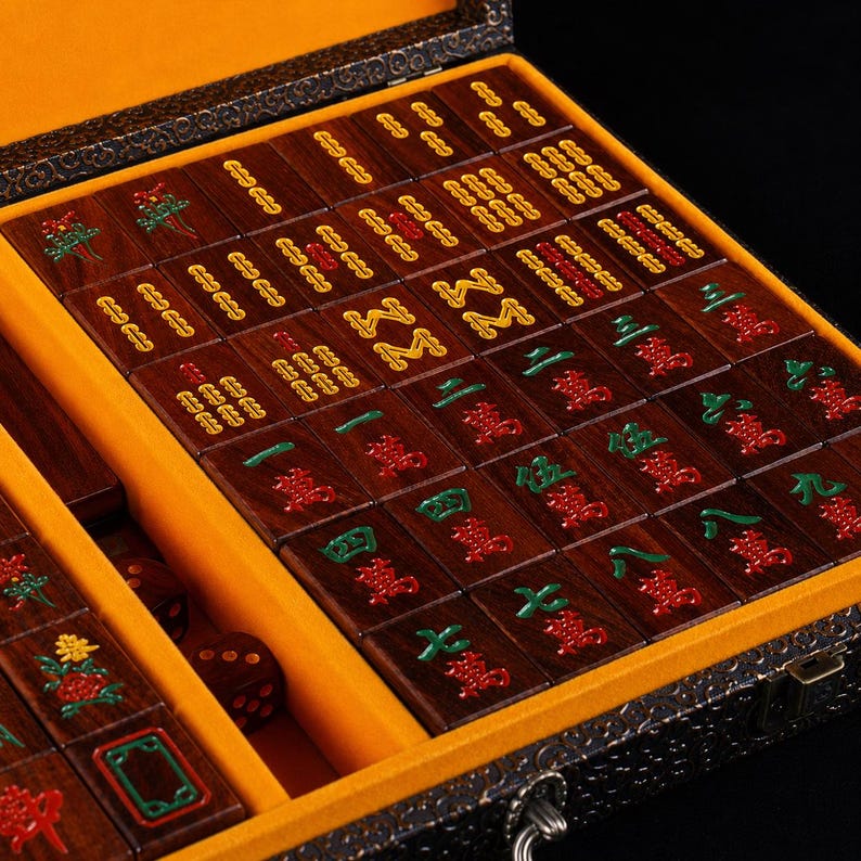 May include: A wooden Mahjong set with tiles in a decorative box. The tiles are dark brown with red, green, and yellow symbols. The box has a yellow interior and a dark exterior with a metal clasp.