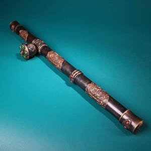 Può includere: Una pipa antica con un corpo in legno scuro e dettagli in metallo ornati. La pipa presenta motivi floreali decorativi, abbellimenti di pietre preziose rosse e verdi e un bocchino in ottone. Il design suggerisce un oggetto vintage o da collezione.