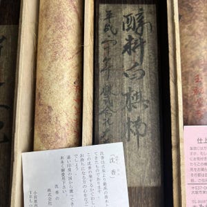 May include: A wooden box containing a rolled paper and a wooden panel with Japanese calligraphy. The paper is beige with a reddish-brown pattern. The wooden panel is dark brown with white ink. A white paper with Japanese text is in the foreground.