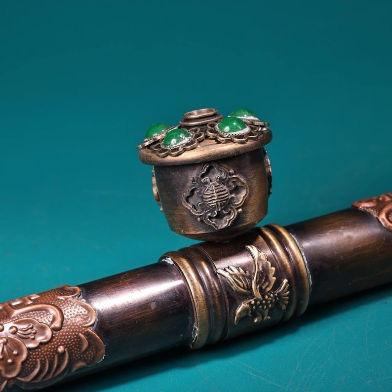 May include: A bronze-colored decorative object with a lid adorned with green gemstones. The object has intricate carvings and is set on a dark brown, ornate rod. The design features floral and bird motifs.
