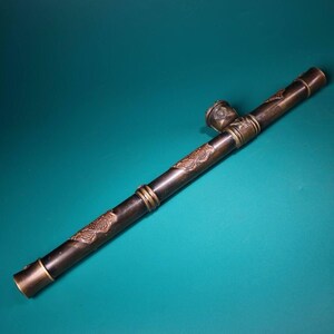 May include: A long, dark brown pipe with ornate bronze-colored accents and decorative floral patterns. The pipe features a bowl and mouthpiece, with brass rings and end caps. The design suggests an antique or vintage style.