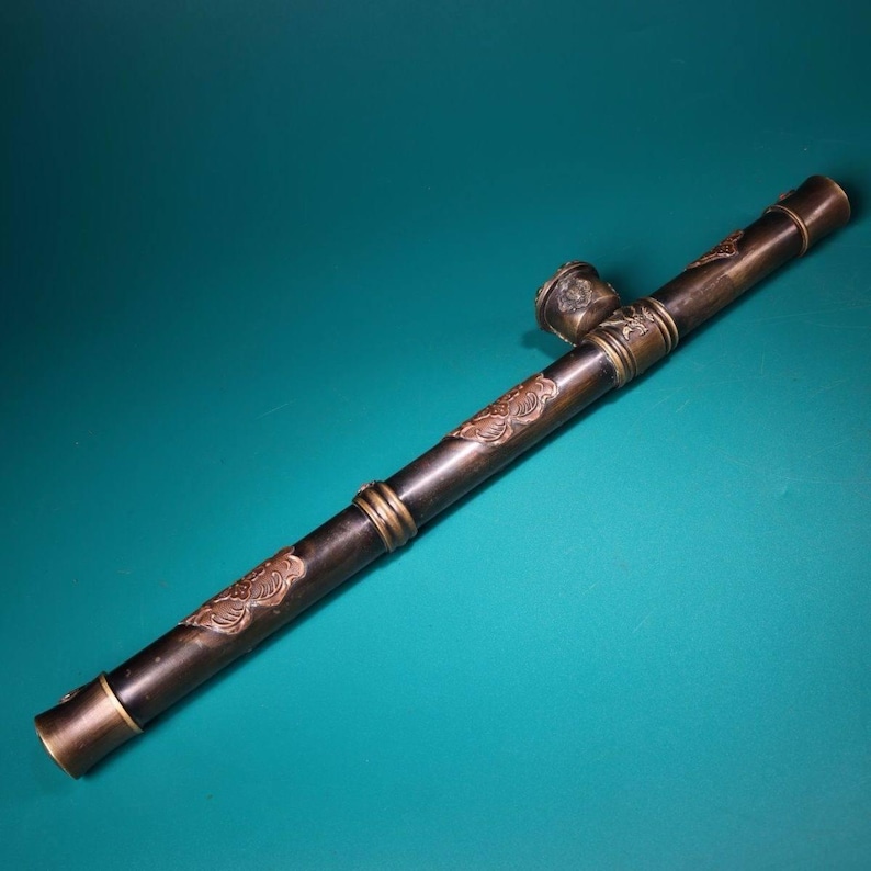 May include: A long, dark brown pipe with ornate bronze-colored accents and decorative floral patterns. The pipe features a bowl and mouthpiece, with brass rings and end caps. The design suggests an antique or vintage style.