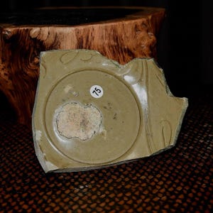 May include: A fragment of a pale green ceramic plate featuring a circular design and the number 75 on a white sticker. The plate has a broken edge and a rough texture. The background is a dark wooden surface.