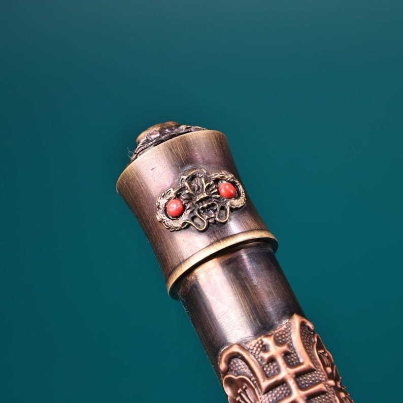 May include: Close-up of a bronze-colored decorative object with intricate carvings and two red gemstone accents. The object has a cylindrical shape with a detailed floral design and a textured surface, set against a teal background.